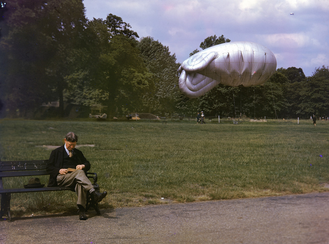 Un hombre lee un libro en un parque de Londres, en el fondo de la fotografía, un globo antiaéreo globo de presa es visible, mientras que un segundo globo se eleva a lo alto en la distancia