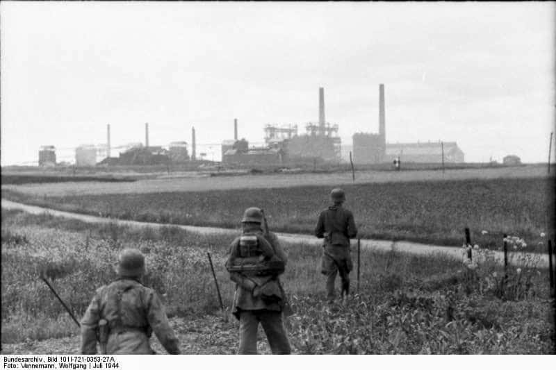 Una patrulla alemana se mueve hacia el área de la fábrica de Colombelles. Algunas de las altas chimeneas utilizadas por los observadores alemanes son visibles en la distancia. Collombelles, julio de 1944