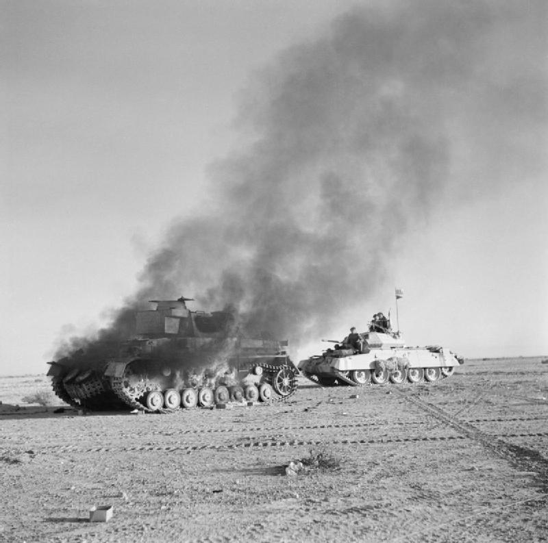 Un blindado alemán destruido durante la batalla, en primer plano, arde mientras que al fondo avanza un tanque Crusader