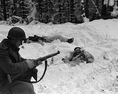 Esta foto, probablemente escenificada, muestra un soldado recargando su fusil ante dos enemigos abatidos. La foto se tomó durante la batalla de las Ardenas.