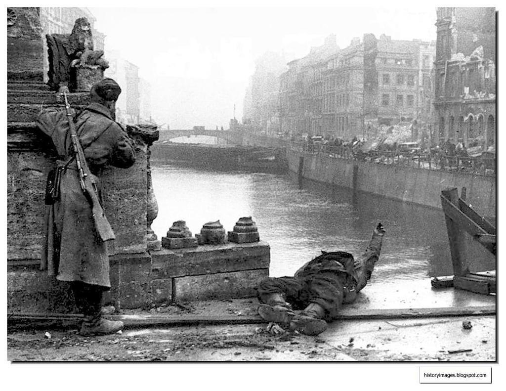 Un ruso se asoma lentamente sobre un puente, mientras a sus pies se encuentra el cadáver de un combatiente alemán